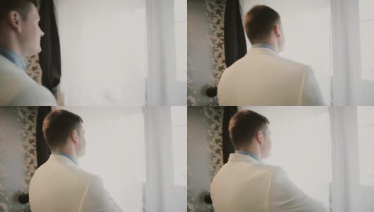 一个穿白色西装的年轻人走近窗户。新郎准备结婚的日子。早上仪式之前高清在线视频素材下载