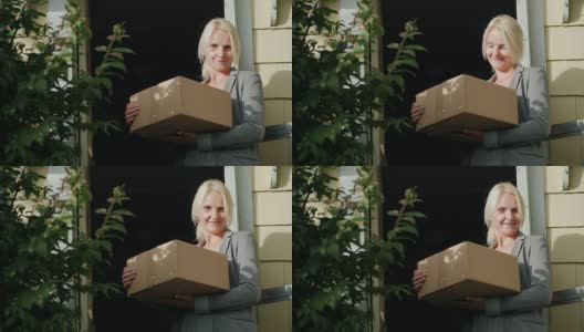 一个拿着包裹的妇女站在她家的门槛上。看着镜头，微笑着。包裹送货上门高清在线视频素材下载