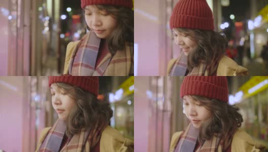 一个亚洲女孩站在商店的橱窗外在一个寒冷的夜晚在城市的圣诞市场，她穿着暖和的衣服。高清在线视频素材下载