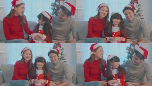 亚洲幸福家庭的肖像戴着圣诞老人的帽子，坐在客厅里。漂亮迷人的父母，穿着红衣服的爸爸妈妈和年幼的女儿看着摄像机准备在家里庆祝圣诞节高清在线视频素材下载