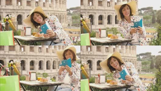 在罗马斗牛场前的一家酒吧餐厅外，一名快乐的年轻游客正在用智能手机自拍，一边喝着咖啡、果汁和玉米片。优雅漂亮的裙子配上大帽子和五颜六色的购物袋高清在线视频素材下载