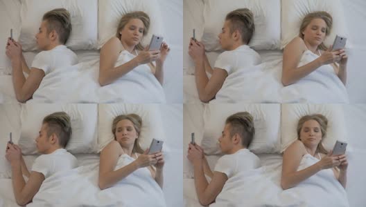 年轻男女背靠背躺在床上用智能手机打字，沉迷于电子产品高清在线视频素材下载