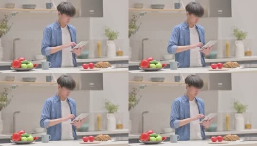 年轻的亚洲男子站在厨房里使用平板电脑高清在线视频素材下载