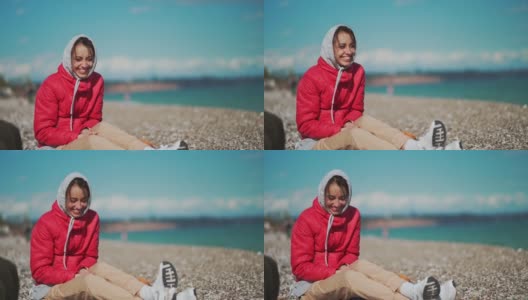 一个穿着红色夹克和连帽衫的年轻女子，在阳光明媚的多风的日子里，坐在卵石海滩上捡卵石高清在线视频素材下载