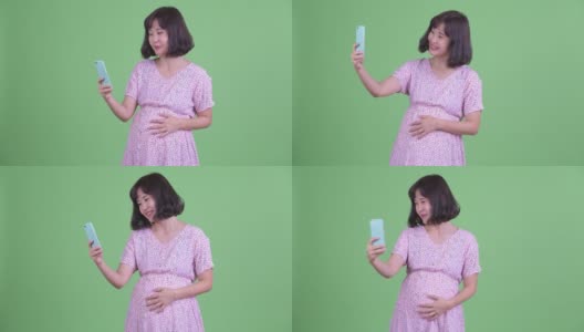 摄影棚拍摄美丽的怀孕亚洲妇女微笑和使用手机在绿色的摄影棚背景高清在线视频素材下载