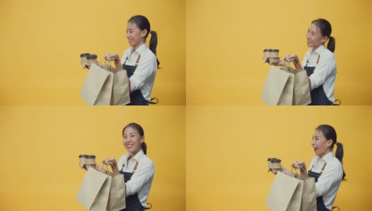 快乐微笑的亚洲女服务员，穿着围裙，友好地看着文案空间的广告，装着咖啡或茶和美味的食物在手工纸袋隔离在黄色的背景上。高清在线视频素材下载