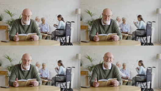 机架上集中了三位老人，两位妇女包括残疾人一人和一位男子，在养老院打牌。老男人戴着眼镜看书，看着相机，笑着坐在桌子旁高清在线视频素材下载