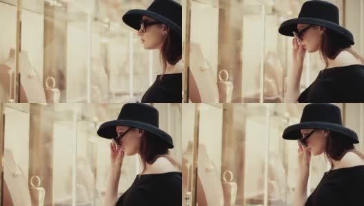 一个漂亮时髦的富家女孩戴着一顶帽子摘下眼镜，在一家奢侈品精品店的橱窗里看商品。慢动作高清在线视频素材下载