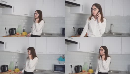 一位戴着眼镜、心情愉快的年轻女子在厨房边吃着食物边讲电话。高清在线视频素材下载