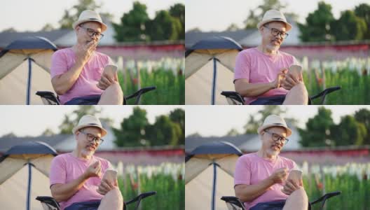 笑容满面的亚洲灰胡子成熟男子坐在户外的花圃里，用手机上的在线约会app聊天。快乐的退休男人穿着粉色衬衫，戴着眼镜，日落时戴着帽子。普通情人节新概念高清在线视频素材下载
