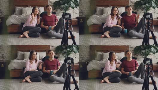 夫妻博主们在录制视频，挥手打手势，有说有笑，他们用三脚架上的相机，坐在卧室地板上。高清在线视频素材下载