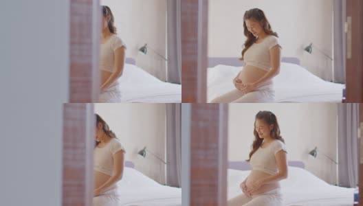 快乐怀孕的亚洲女人坐在床上抱着和抚摸着自己的大肚子，年轻女人的怀孕享受和未来的生活放松在家里，母亲和怀孕的概念，柔软的焦点高清在线视频素材下载
