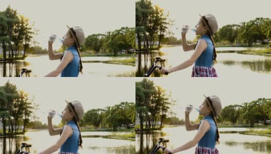 一个小女孩戴着帽子在城市公园的河边喝水，在夏天的夕阳下，坐在一辆自行车上。肖像。的处理。4 k。高清在线视频素材下载