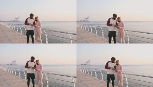 快乐的年轻夫妇戴着防护面具在美丽的日出时在海边使用手机，慢镜头高清在线视频素材下载