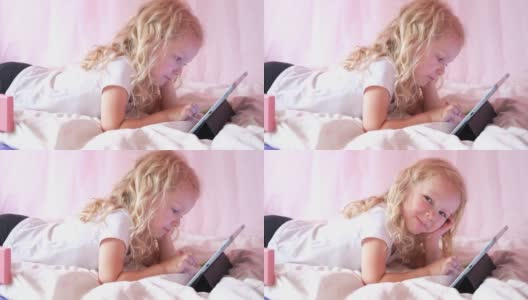 高加索好奇卷曲可爱的学龄前女孩使用数字平板技术设备躺在床上独自。小孩子拿着垫子在家里上网玩游戏。儿童科技上瘾高清在线视频素材下载