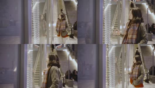 在一个寒冷的夜晚，一个亚洲女孩站在城市商店的橱窗外，她穿着暖和的衣服。高清在线视频素材下载